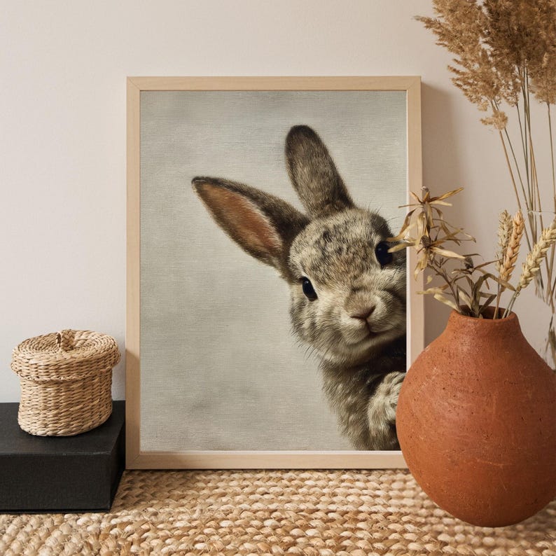May include: A framed print featuring a detailed illustration of a rabbit peeking from the side. The rabbit has gray and brown fur, with long ears and dark eyes. The artwork is set against a neutral background, and the frame is light-colored.