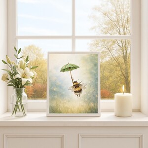 K&ouml;nnte beinhalten: Ein gerahmter Kunstdruck mit einer Illustration einer Hummel, die einen gr&uuml;nen Regenschirm mit wei&szlig;en Punkten h&auml;lt und in einer vertr&auml;umten, sonnendurchfluteten Wiese schwebt. Das Kunstwerk steht auf einer Fensterbank neben einer Vase mit wei&szlig;en Blumen und einer brennenden Kerze.