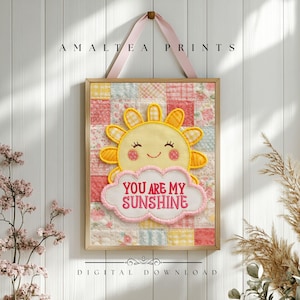 Könnte beinhalten: Gerahmter Wandkunstdruck mit Patchwork-Hintergrund. Das Kunstwerk zeigt eine lächelnde gelbe Sonne mit rosa Wangen und den Schriftzug "YOU ARE MY SUNSHINE" in rosa Stickerei. Der Druck wird von einem rosa Band gehalten.