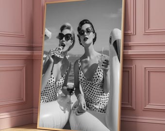 Puede incluir: Impresión enmarcada en blanco y negro de dos mujeres con trajes de baño de lunares y gafas de sol, disfrutando de pizza y bebidas en cisnes inflables en la playa. La imagen tiene una estética vintage.