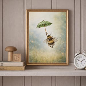 K&ouml;nnte beinhalten: Ein gerahmter Kunstdruck zeigt eine skurrile Illustration einer Hummel, die einen gr&uuml;nen Regenschirm mit wei&szlig;en Punkten h&auml;lt und &uuml;ber einem Feld mit Wildblumen schwebt. Das Kunstwerk ist in einem goldenen Rahmen gefasst und verleiht der naturnahen Szene einen Hauch von Eleganz.
