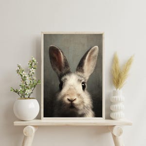 Op de afbeelding: Een ingelijste print met een close-up van een konijnengezicht. Het konijn heeft grijs en wit haar, grote oren en donkere ogen. Het kunstwerk wordt getoond op een lichtgekleurde houten plank met decoratieve vazen en planten.