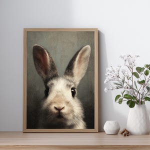 Op de afbeelding: Een ingelijste print met een close-up van het gezicht van een konijn. Het konijn heeft grijs en wit haar, grote oren en donkere ogen. Het kunstwerk staat in een houten lijst en wordt op een houten oppervlak tentoongesteld.
