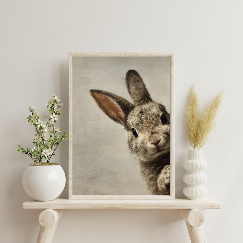May include: A framed print featuring a close-up of a curious rabbit. The rabbit has brown and gray fur, with long ears and dark eyes. The artwork is displayed on a light-colored wooden shelf, with decorative vases and plants.
