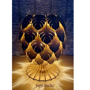 May include: A decorative wooden table lamp with a leaf-shaped shade. The lamp features a base with a scalloped design and a shade composed of layered, dark-colored leaves. The lamp is illuminated from within, casting a warm glow. The text "yuyyi studio" is visible.