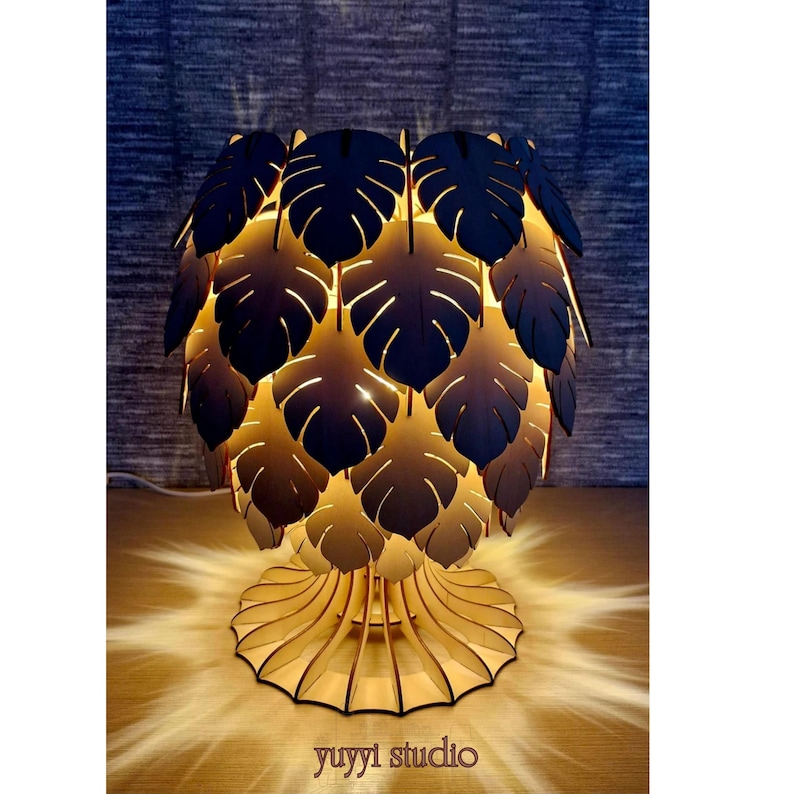 May include: A decorative wooden table lamp with a leaf-shaped shade. The lamp features a base with a scalloped design and a shade composed of layered, dark-colored leaves. The lamp is illuminated from within, casting a warm glow. The text "yuyyi studio" is visible.