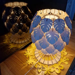 May include: A decorative wooden lamp with a leaf design. The lamp has a white base and a shade with blue and white leaf cutouts. The lamp is sitting on a wooden surface with a yellow floral design. The text "yuyyi studio" is visible.