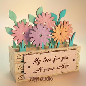 Peut inclure: Bo&icirc;te &agrave; fleurs en bois avec des fleurs d&eacute;coup&eacute;es au laser roses, violettes et oranges. La bo&icirc;te porte une pancarte avec l'inscription "My love for you will never wither" et le texte "yuyyi studio". Les fleurs ont des feuilles et de l'herbe vertes.