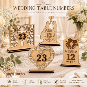 May include: Four laser-cut wooden wedding table numbers with stands. Designs include a tree with a heart, a couple holding a heart, a heart with flower borders, and a couple silhouette. Text includes 'Table 12' and the number '23'.