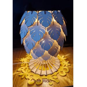 May include: A wooden table lamp with a leaf design. The lamp shade is made of layered, light-colored wooden leaves. The base is a yellow, floral-shaped design. The lamp is lit, casting a warm glow. The text "yuyyi studio" is visible on the base.