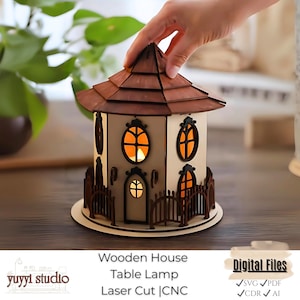 Pode incluir: Uma luminária de mesa em forma de casa de madeira, com telhado marrom e corpo bege. A luminária tem janelas ovais e uma pequena cerca ao redor da base. O texto "Wooden House Table Lamp Laser Cut | CNC" é visível.