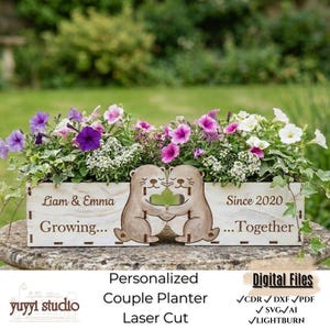 May include: A wooden planter box with laser-cut otters holding hands, filled with colorful petunias. The planter includes the text "Liam & Emma Growing... Since 2020 ...Together". A personalized couple planter.