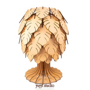 May include: A wooden lamp shaped like a tree, featuring a base, trunk, and a canopy of leaf-shaped cutouts. The lamp is made of light-colored wood and has a natural, organic aesthetic. The text "yuyyl studio" is visible at the base.