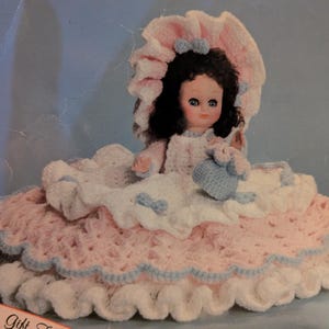 Peut inclure: Poupée faite à la main avec une robe crochetée rose et blanche, bordée de bleu clair. La poupée a des cheveux foncés et porte un bonnet assorti. Elle tient un petit sac à main crocheté bleu.