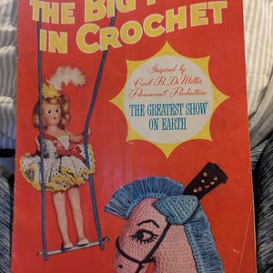 May include: A vintage crochet pattern book titled "The Big Top in Crochet" with a red cover. The cover features a doll on a swing, a pink crocheted horse, and text. The book is inspired by Cecil B. DeMilles Paramount Production.