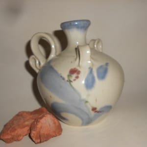 May include: A cream-coloured ceramic vase with blue and red floral patterns. It has two small handles and a larger, curved handle. The rim is painted blue. Two reddish-brown rocks are in the foreground. The vase is likely handmade.