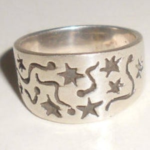 May include: A silver ring with a wide band featuring a cut-out design of stars and wavy lines.