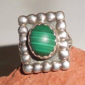 May include: A silver ring with a green malachite stone set in a square bezel. The ring is decorated with a beaded border.