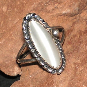May include: A silver ring with a white oval-shaped stone set in a rope-like design. The ring is on a brown rock.