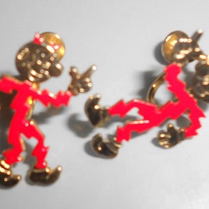 Może przedstawiać: Para złotych kolczyków z czerwonymi figurkami diabłów. Figurki są stylizowane i mają kreskówkowy wygląd. Kolczyki są zaprojektowane z tyłem w kształcie motyla.