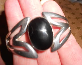 May include: A silver cuff bracelet with a black oval gemstone set in the center. The bracelet has a geometric design with openwork details.