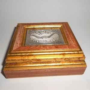 Op de afbeelding: Een kleine houten doos met een gouden frame en een zilverkleurig reli&euml;fdesign op de deksel. Het ontwerp toont een vogel in vlucht met bloemmotieven.