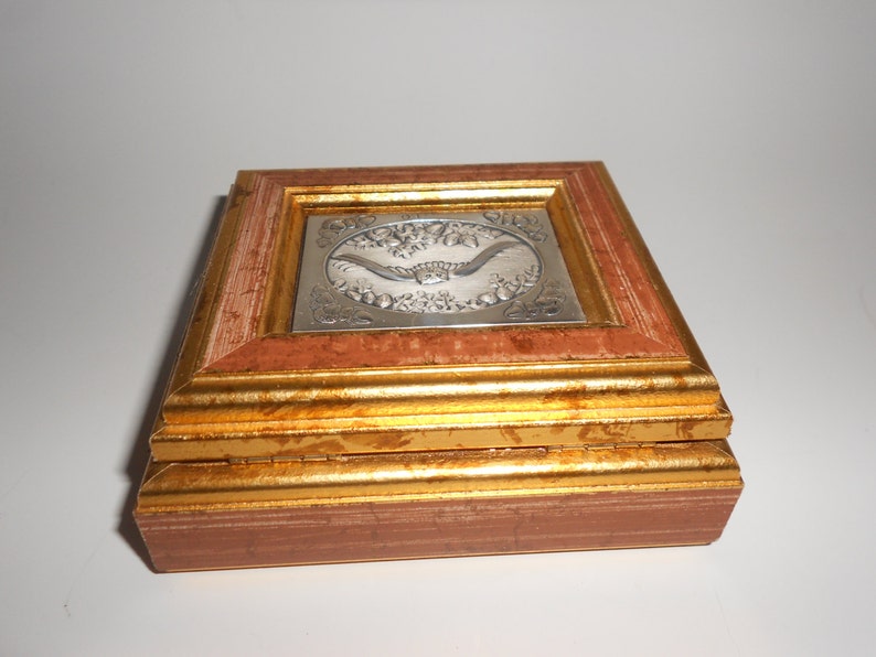 Op de afbeelding: Een kleine houten doos met een gouden frame en een zilverkleurig reli&euml;fdesign op de deksel. Het ontwerp toont een vogel in vlucht met bloemmotieven.