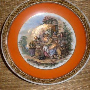 Puede incluir: Un plato decorativo con un borde naranja y una escena central que representa figuras en un entorno rural. El plato presenta un borde dorado y una ilustración detallada, mostrando una estética vintage.
