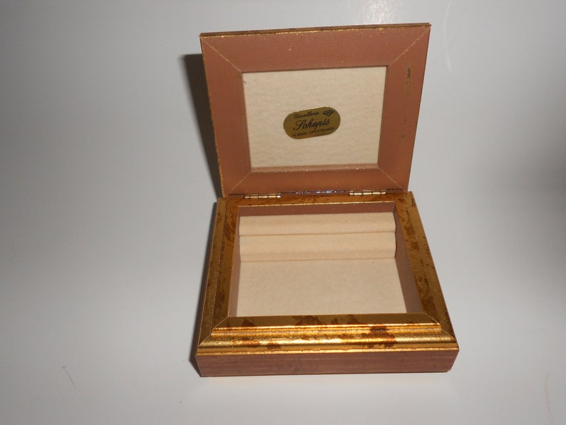 Op de afbeelding: Een klein houten doosje met een gouden rand en een fluwelen interieur. Het doosje is open en het deksel is opengeklapt. Het deksel heeft een label met de tekst "Creations of Shepnis".