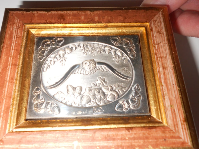 Op de afbeelding: Een zilveren ingelijst beeld met een reli&euml;fsculptuur van een uil die over een boslandschap met eikels vliegt. De sculptuur is gesigneerd met "Eo."