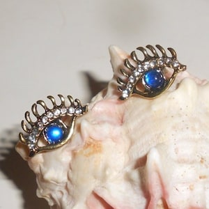 May include: A pair of gold-tone earrings with blue gemstone eyes and clear rhinestone accents. The earrings are shaped like an eye with a long, curved lash.