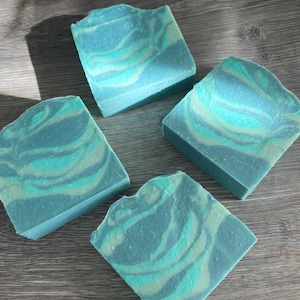 May include: Four square bars of handmade soap with a marbled design. The soap features shades of blue and green, creating a swirling pattern. The bars are arranged on a wooden surface.