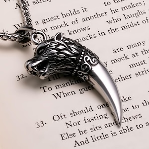 May include: A silver-colored pendant necklace featuring a wolf head design with a large, pointed tooth. The wolf head has detailed fur and facial features. The necklace is on a chain, and the background is a book page.