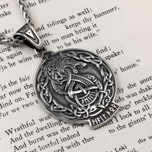 May include: A silver-colored pendant necklace featuring a detailed wolf design within a circular frame of Celtic knots. The pendant includes runes and is suspended from a matching chain, set against a backdrop of printed text.