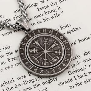 May include: A silver-toned pendant necklace featuring a circular pendant with a Viking compass design. The pendant has runic symbols around the edge and is suspended from a chain. The necklace rests on a book page.