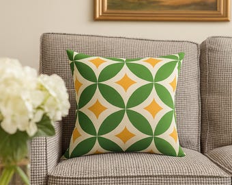 Op de afbeelding: Een vierkant decoratief kussen met een geometrisch patroon van groene overlappende cirkels en gele ruitvormen. Het kussen ligt op een grijs en wit patroonbank, met een schilderij op de achtergrond.