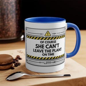 Puede incluir: Taza de cerámica blanca con interior y asa azules. La taza presenta el texto "OF COURSE SHE CAN'T LEAVE THE PLANT ON TIME" dentro de un diseño gris y amarillo con rayas de peligro. Una cuchara y un pastelito están en primer plano.
