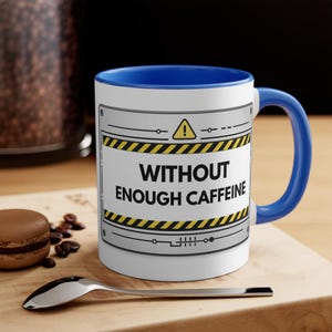 May include: White ceramic mug with a blue interior and handle. The mug features the text "WITHOUT ENOUGH CAFFEINE" in black, with a yellow and black hazard stripe design. A spoon and a macaron are in the foreground.