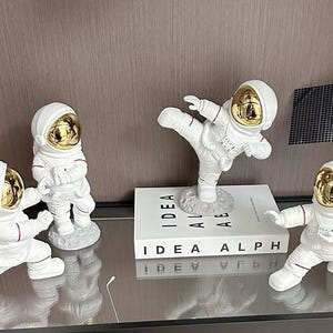 Könnte beinhalten: Eine Sammlung weißer Astronautenfiguren mit goldenen Visieren und roten Akzenten. Eine Figur befindet sich in einer Kick-Pose, balancierend auf einem Buch mit der Aufschrift "IDEA ALPH". Ein Modell eines Satelliten ist ebenfalls vorhanden.
