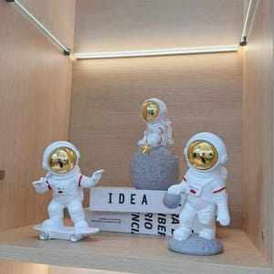 May include: Three astronaut figurines in white suits with gold visors. One astronaut is on a skateboard, another is holding a small ball, and the third is seated on a gray moon with a dangling star. The word "IDEA" is visible on a book.