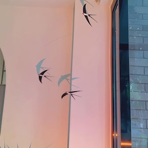 Könnte beinhalten: Wanddekoration mit stilisierten schwarzen Schwalben im Flug vor einer hellrosa Wand. Die Vögel mit ausgebreiteten Flügeln erzeugen ein Gefühl der Bewegung. Das Design fügt dem Raum einen Hauch von naturnaher Dekoration mit einem dezenten Farbverlauf hinzu.