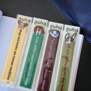 Puede incluir: Cuatro marcapáginas coloridos con temática de perros, con caras de perros bordadas y textos humorísticos. Los marcapáginas son de color amarillo, verde, marrón y verde oliva. Los textos incluyen "dogs and books are human medicine" y "drink coffee read books pet dogs".