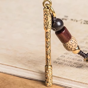 May include: A gold-colored metal pendant with intricate carvings and Chinese characters. The pendant is attached to a beaded strand with wooden and dark beads. The background is a book with Chinese text.