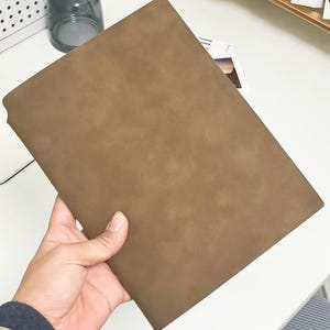 Puede incluir: Un cuaderno rectangular marrón con una superficie texturizada. El cuaderno se sostiene en una mano, mostrando su tamaño y material. La cubierta parece estar hecha de un material similar al ante.