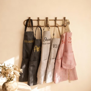May include: Five striped aprons with adjustable neck straps and front pockets hang from a wooden wall rack. The aprons feature embroidered names including Amanda, Ava, Jess, and Eli, with varying color schemes of black, grey, and red stripes.