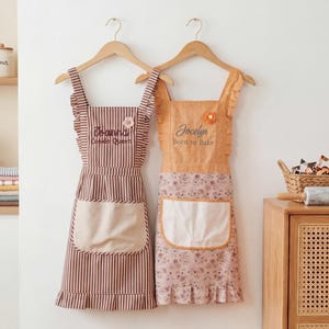 May include: Two aprons hanging on wooden hangers. The left apron features brown and white vertical stripes, a beige pocket, and the text "Joanna Cookin Queen". The right apron is solid orange with a floral skirt, white pocket, and "Jocelyn Born to Bake".