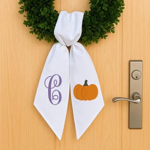 May include: White door sash with a purple embroidered letter "C" and an orange pumpkin design. The sash is hanging on a green wreath, perfect for autumn decor. The door has a silver handle.