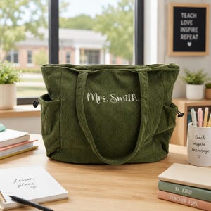 May include: A dark olive green corduroy tote bag featuring two side pockets with black drawstrings and the embroidered white text Mrs. Smith centred on the front. The bag has two matching fabric shoulder straps and a zippered top closure.