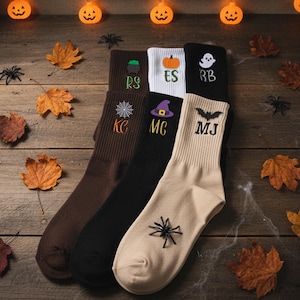 May include: Six pairs of Halloween socks in shades of brown, black, white, and beige. Each sock has a different design, including a spider web, pumpkin, ghost, witch's hat, and spider. The socks are displayed on a wooden surface with decorative leaves and spider decorations.