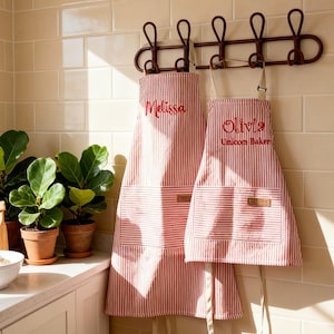 May include: Two red and white striped aprons with beige neck straps and waist ties. The larger apron features the embroidered name Melissa in red, while the smaller apron displays the name Olivia and the text Unicom Baker in red embroidery.